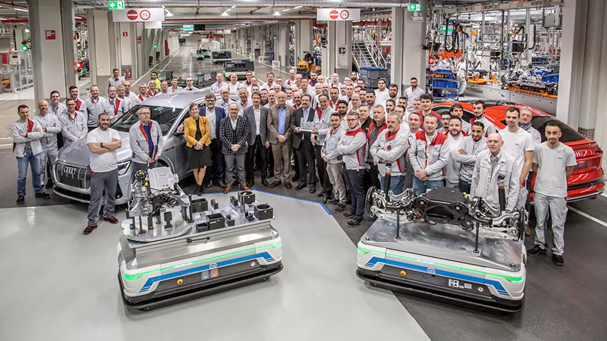 A group photo of blue collar workers and management standing around a red and grey Audi e-tron, along with two self-driving transport systems